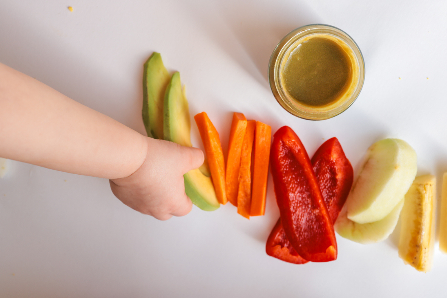 A child's hands reaching for freshly chopped vegetables on a white surface. Includes diced avocado, carrot, apple slices, bell pepper, and tomato pieces. Minimalist food composition