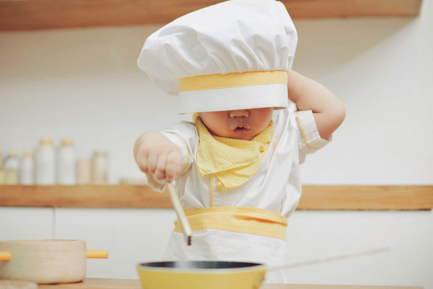 Baby in chef hat mixing something in bowl in white kitchen. 