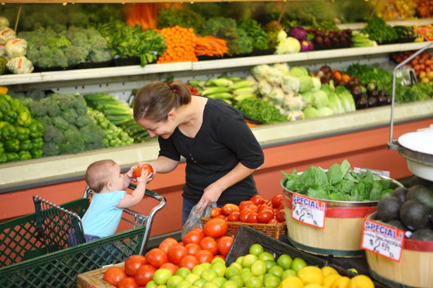 Woman shopping in produce section with baby in cart, woman handing baby a tomato. 