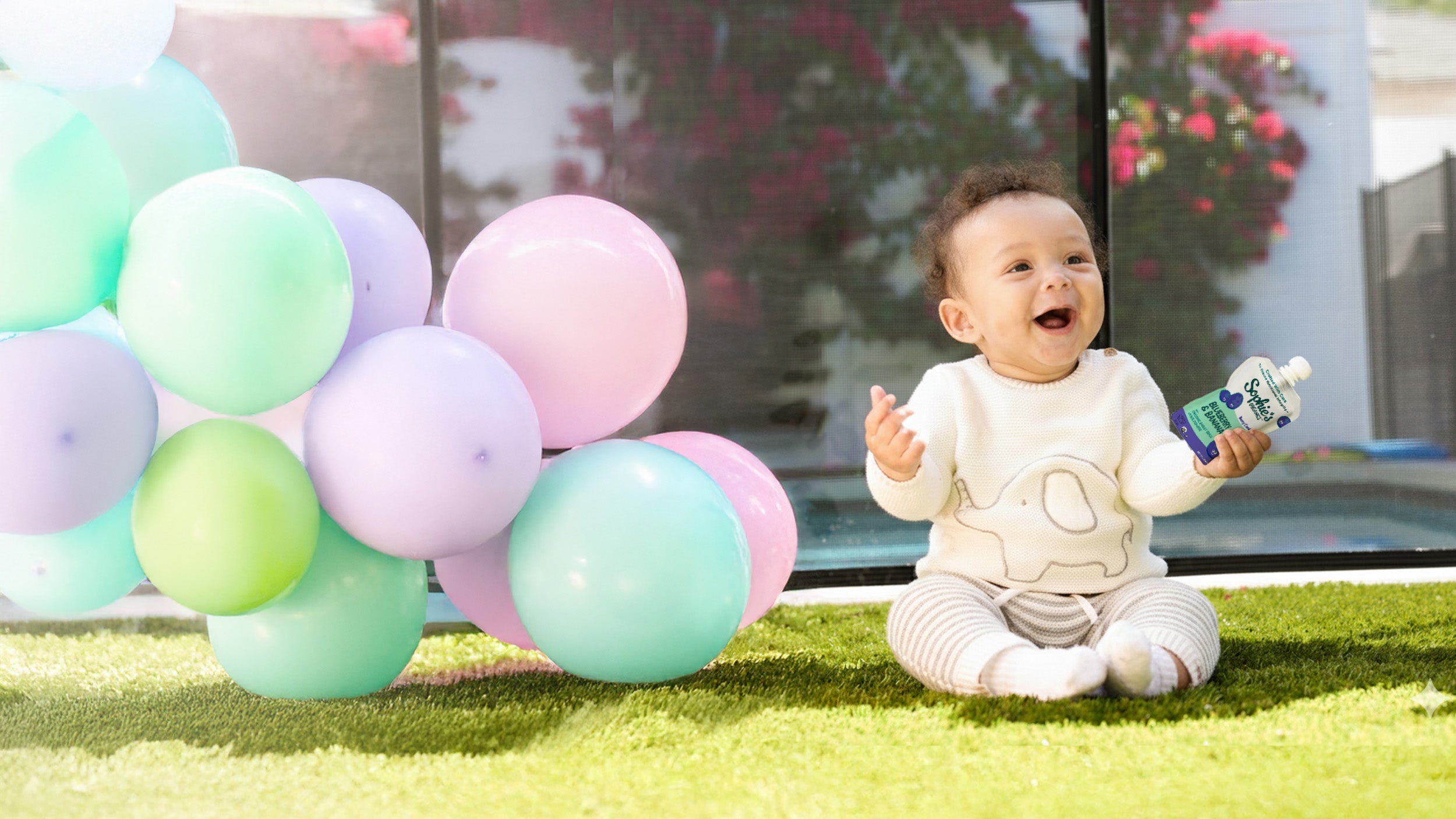 Happy baby holding Sophie's Organics Berry Cool organic baby food pouch outdoors with balloons. 