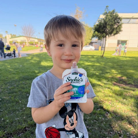 Child smiling with Sophie's Organics organic baby food pouch at the playground.