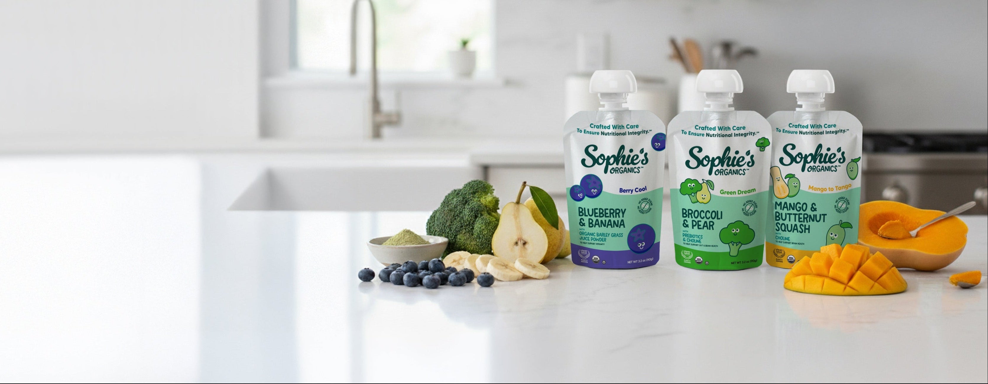 Three pouches of Sophie's Organic baby food on a kitchen counter with fruits and vegetables.